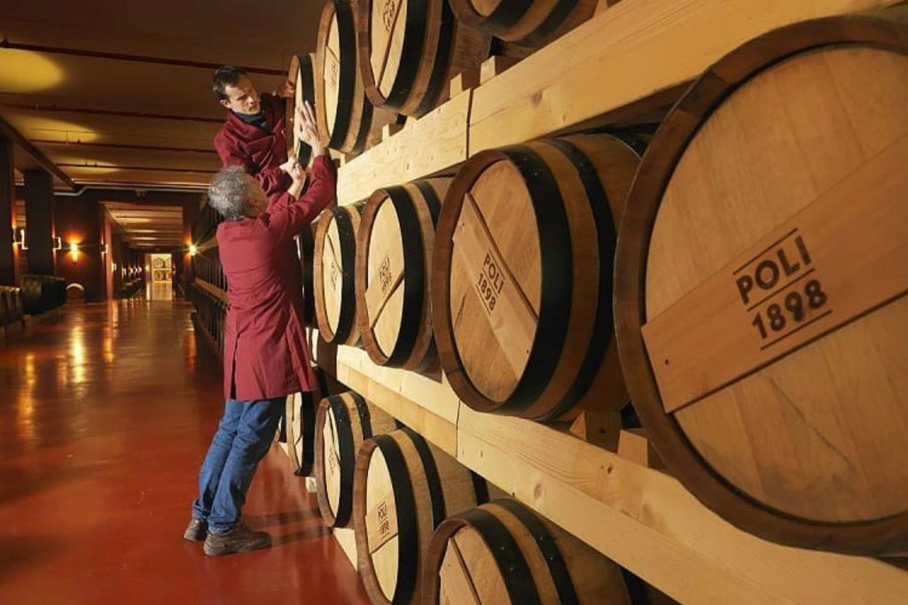 빨간 코트를 입은 두 사람이 빨간색 바닥이 있는 창고의 선반에 쌓인 커다란 나무 통을 검사하고 있습니다.