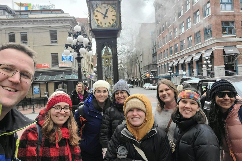 건물과 가로등이 보이는 도시 거리에 있는 개스타운 증기 시계 앞에서 한 무리의 사람들이 미소를 짓고 있습니다.