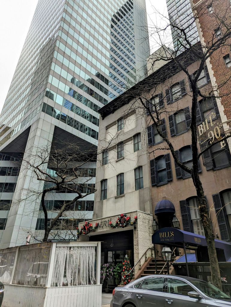 현대적인 유리 마천루와 오래된 벽돌 건물이 있는 도시 거리 풍경 ('Bill's 90 E.' 라고 적힌 간판 포함)