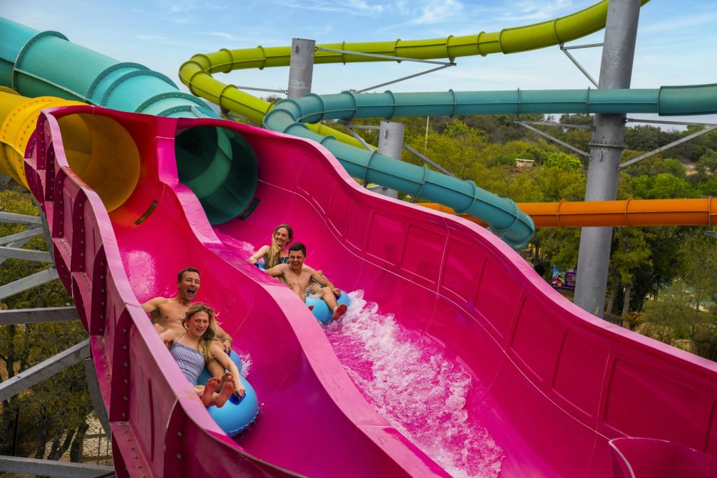 파란색 공기 주입식 미끄럼틀을 탄 4명이 노란색과 녹색 미끄럼틀로 둘러싸인 밝은 분홍색 물 미끄럼틀 아래로 미끄러집니다.