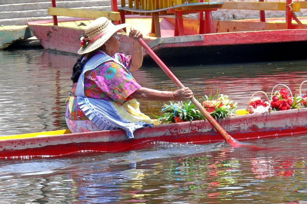 알록달록한 색상의 드레스와 모자를 쓴 여성이 붉은 보트를 타고 잔잔한 물가에서 다른 보트와 함께 노를 젓고 있습니다.