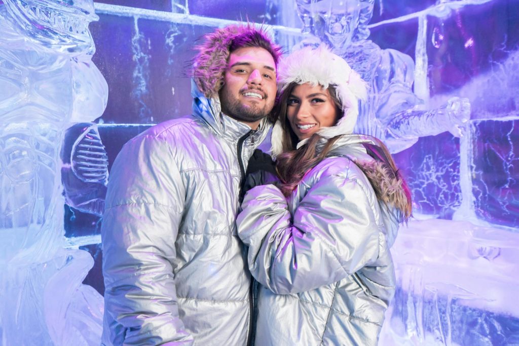 은색 파카를 입은 커플이 보라색 조명이 있는 정교한 얼음 조각상 앞에 서서 웃고 있습니다.