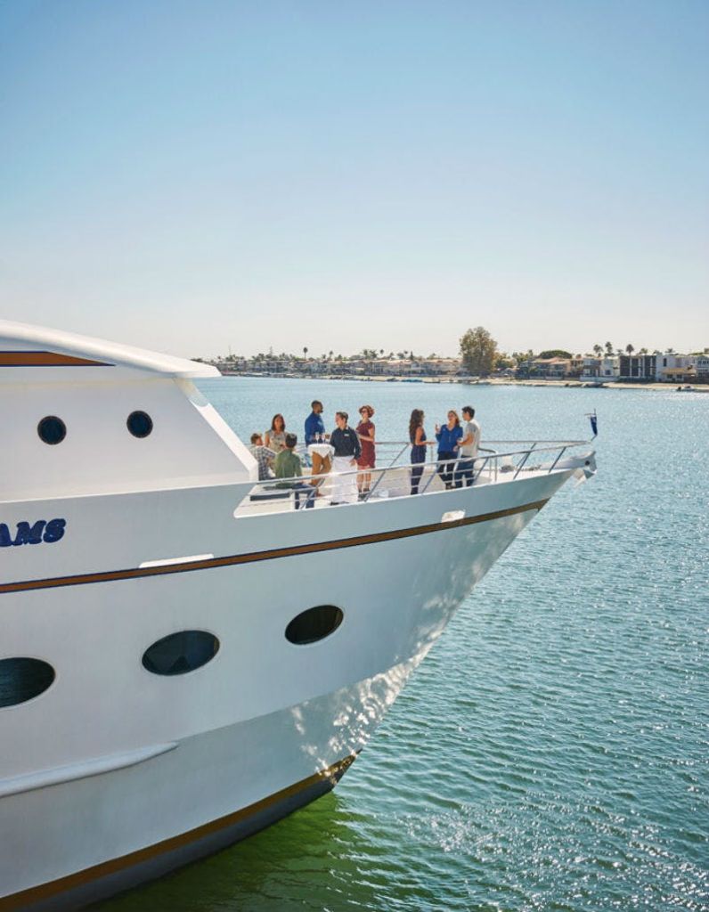 맑은 하늘 아래 먼 해안선이 보이는 잔잔한 물가에서 사람들이 하얀 요트의 갑판에 모여 있습니다.