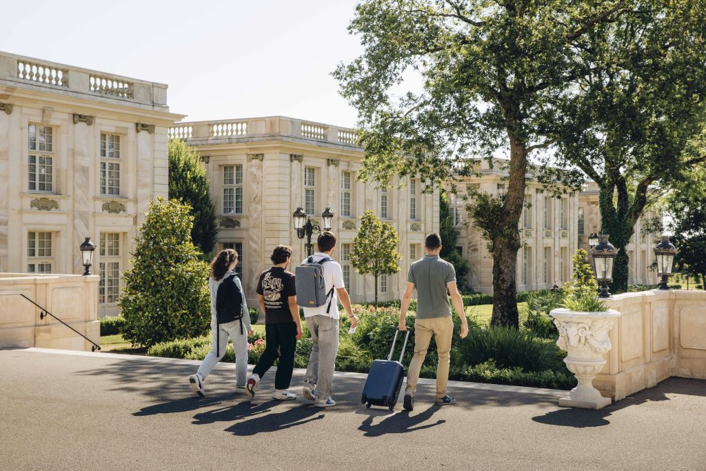 화창한 날 나무와 녹지로 둘러싸인 짐을 들고 고전적인 스타일의 건물을 향해 걸어가는 네 명의 사람들.