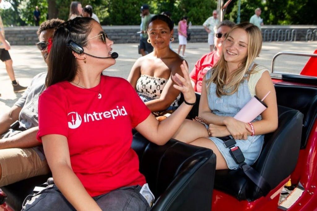 투어 차량에 앉은 일행 중 한 명은 마이크를 착용하고 말하고, 다른 한 명은 미소를 지으며 경청합니다.