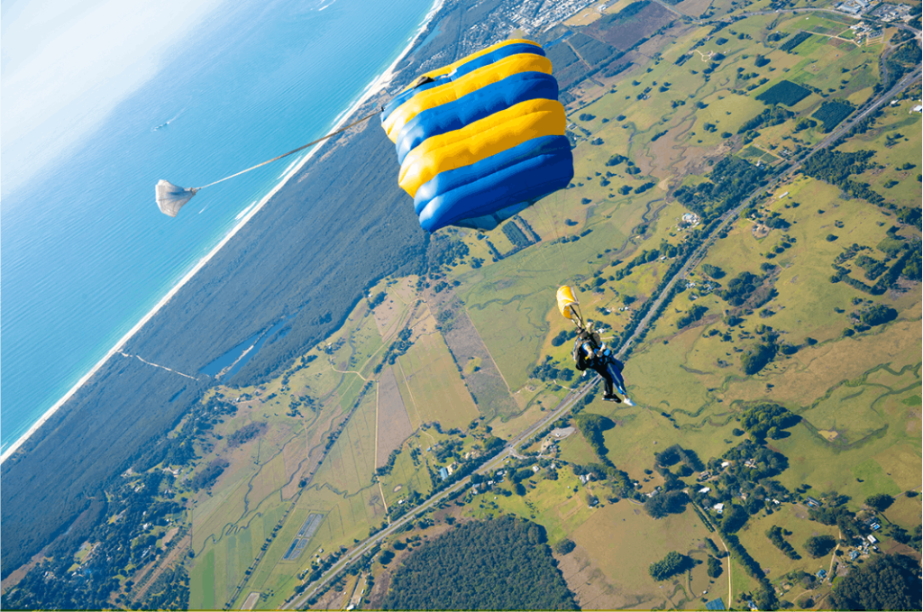 한 사람이 밭, 나무, 먼 바다가 있는 해안 풍경을 보며 낙하산을 타고 있습니다. 낙하산은 파란색과 노란색입니다.