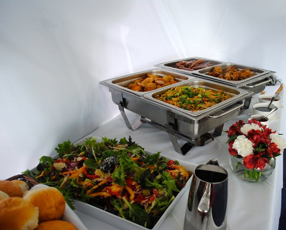 다양한 핫푸드의 차핑 요리, 샐러드, 빵 롤, 피처, 빨간색과 흰색 꽃의 중심이 되는 뷔페 세트입니다.