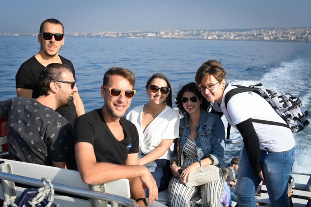 선글라스를 끼고 보트에 앉아 도시의 스카이라인을 배경으로 웃고 있는 6명의 사람들.