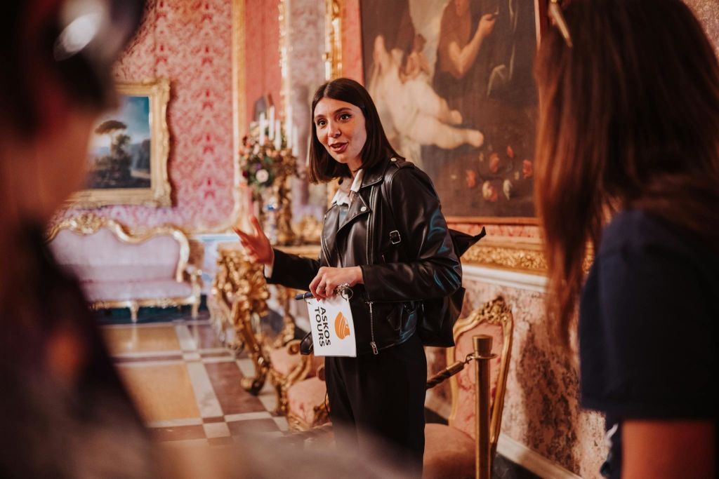 안내 책자를 들고 있는 가죽 재킷을 입은 투어 가이드가 그림과 앤티크 가구가 있는 화려한 방에 서 있습니다.