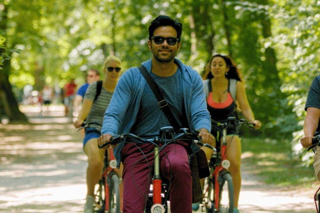 한 무리의 사람들이 화창한 날 나무가 우거진 길에서 자전거를 타고 있습니다. 앞에 있는 사람은 선글라스와 파란색 후드티를 착용합니다.