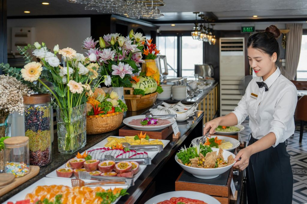 정장 차림의 여성은 조명이 밝은 식사 공간에서 다양한 요리, 꽃, 과일 바구니가 있는 뷔페의 음식을 제공합니다.