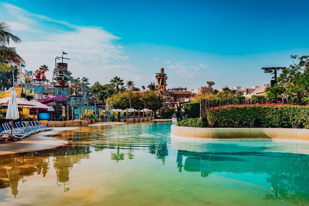 미끄럼틀, 한가로운 강, 열대 식물, 맑고 푸른 하늘 아래 구조물이 있는 워터파크.