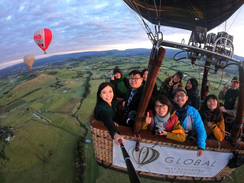 경치 좋은 풍경 위로 높이 솟은 "Global Ballooning Australia" 배너가 달린 열기구 바구니에 담긴 미소 짓는 사람들.