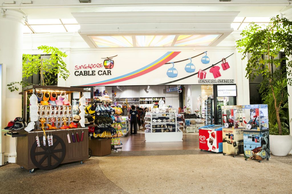 '싱가포르 케이블카' 라고 적힌 선물 가게 입구에는 모자, 장난감, 옷 등의 기념품이 전시되어 있습니다. 안에는 두 사람이 서 있습니다.