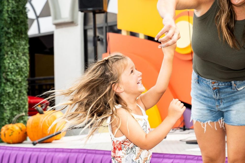 긴 머리를 한 어린 소녀가 어른과 손을 잡고 웃으며 춤을 추고 있습니다. 밝고 다채로운 배경과 캐주얼한 분위기.