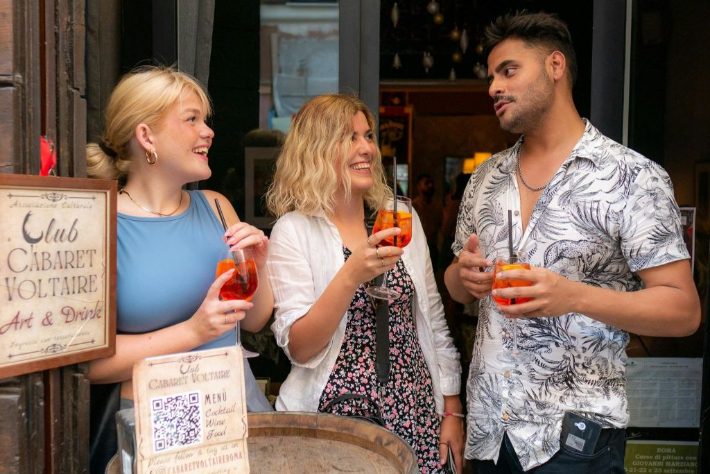 세 사람이 공연장 밖에 서서 미소를 짓고 빨간색 음료가 담긴 잔을 들고 있습니다. 메뉴가 통에 표시됩니다.