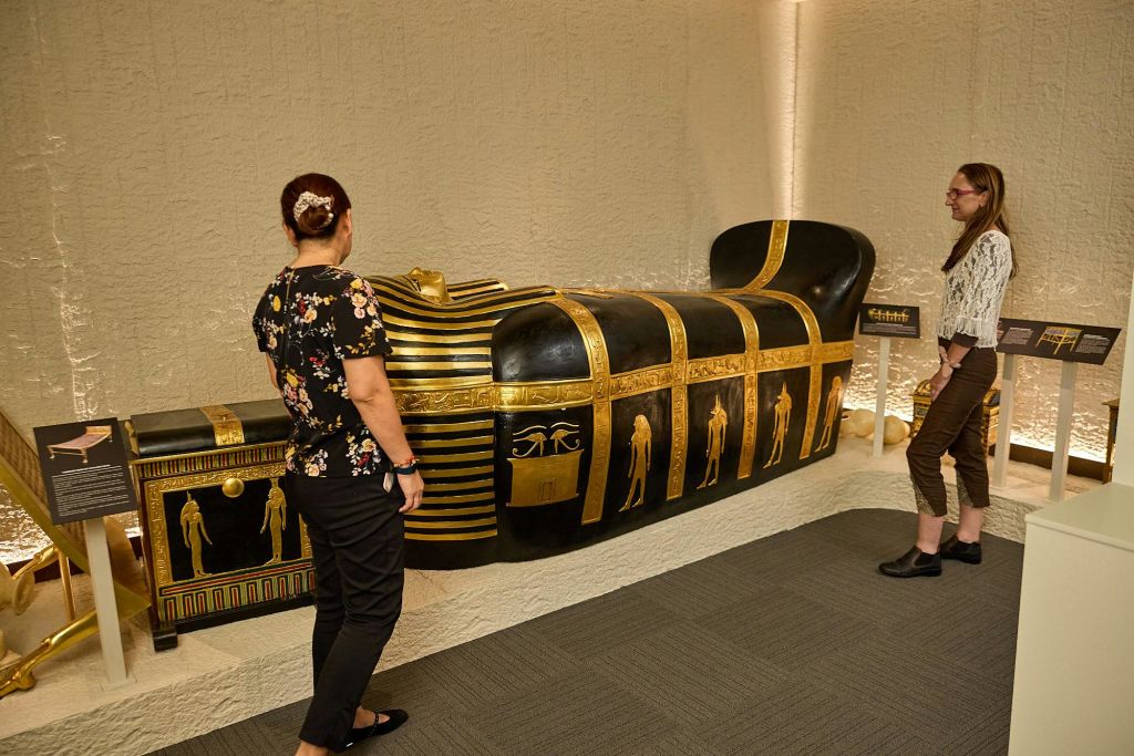 박물관 전시관에서 두 사람이 검은색과 금색 석관 근처에 서 있습니다. 정보 명판은 그 옆에 표시됩니다.