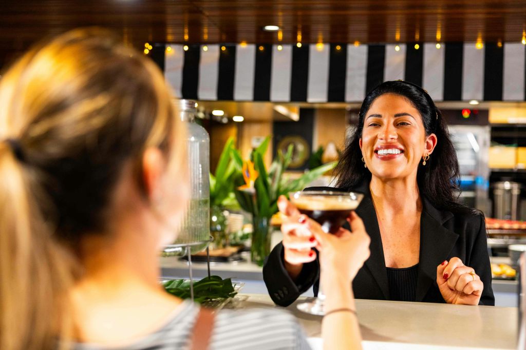 검은색 블레이저를 입은 여성이 화분과 줄무늬 장식이 배경인 레스토랑에서 누군가에게 음료를 건네며 미소를 짓고 있습니다.