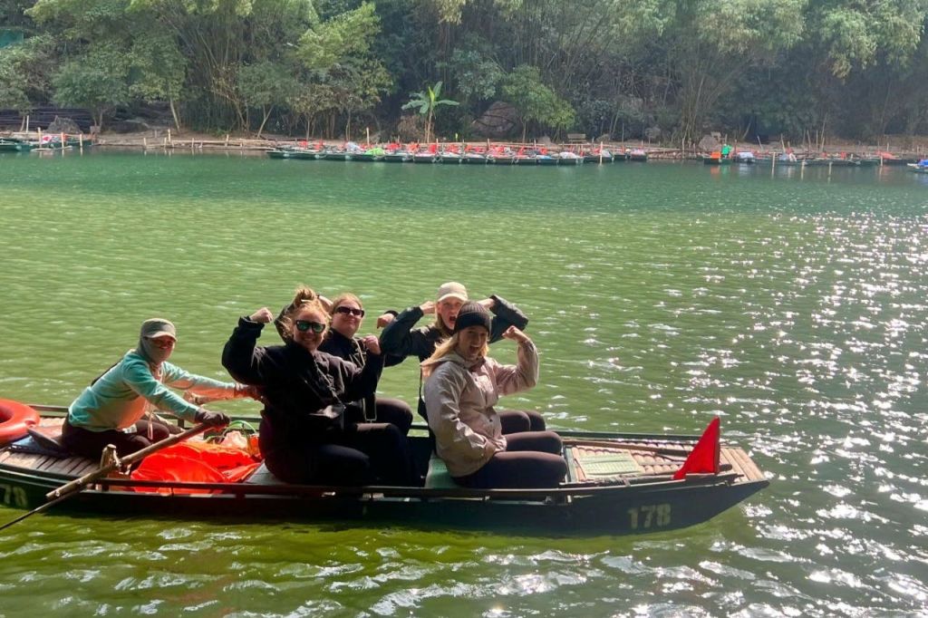 나무와 다른 보트를 배경으로 푸른 강가의 작은 보트에 네 명이 앉아 있습니다.