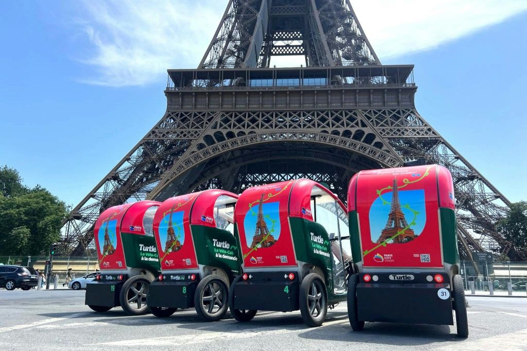 4대의 소형, 적색 및 녹색 전기 차량이 맑은 날 에펠탑 앞에 나란히 주차되어 있습니다.