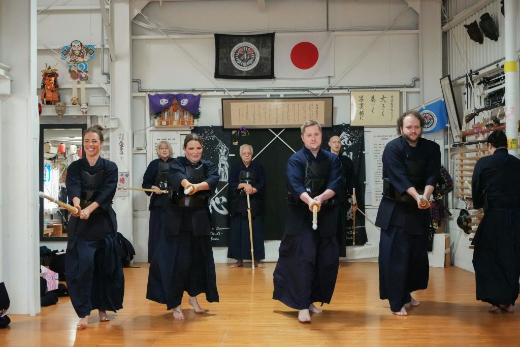 전통 무술 복장을 한 사람들이 도장에서 목검을 들고 현수막과 깃발을 배경으로 연습하고 있습니다.