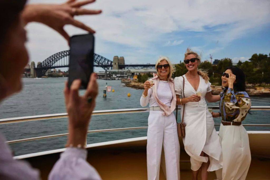 화창한 날 시드니 하버 브리지를 배경으로 세 명의 여성이 보트에서 미소를 짓고 음료를 마시고 있습니다.