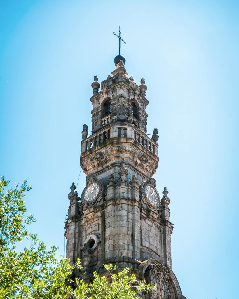 청명한 푸른 하늘 아래 화려한 디테일이 돋보이는 역사적인 석조 시계탑으로, 전면에 나뭇가지가 보입니다.