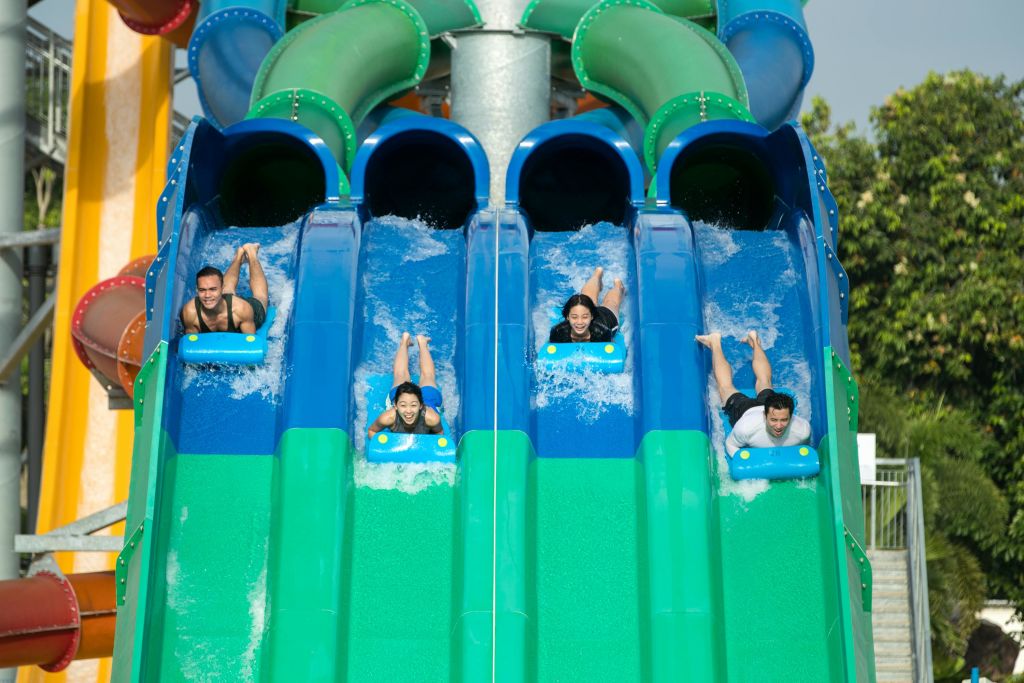 4명이 평행한 녹색과 파란색 워터 슬라이드를 미끄러지며 미소를 짓고 매트를 붙잡고 있습니다. 나무는 배경에 있습니다.