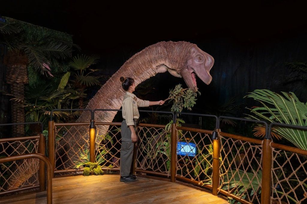 식물과 조명이 있는 테마 전시 환경에서 현실적인 공룡 모델 근처에서 단풍을 들고 있는 유니폼을 입은 사람.