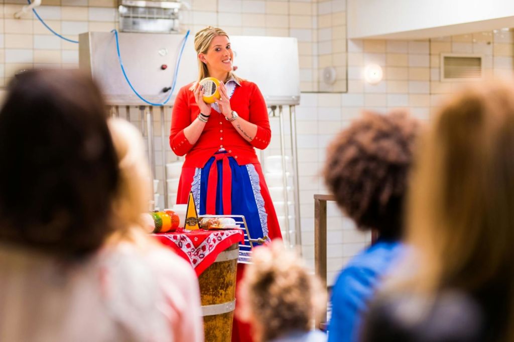 빨간색과 파란색 옷을 입은 여성이 타일로 덮인 방에서 소규모 청중에게 프레젠테이션을 하는 동안 치즈를 들고 있습니다.