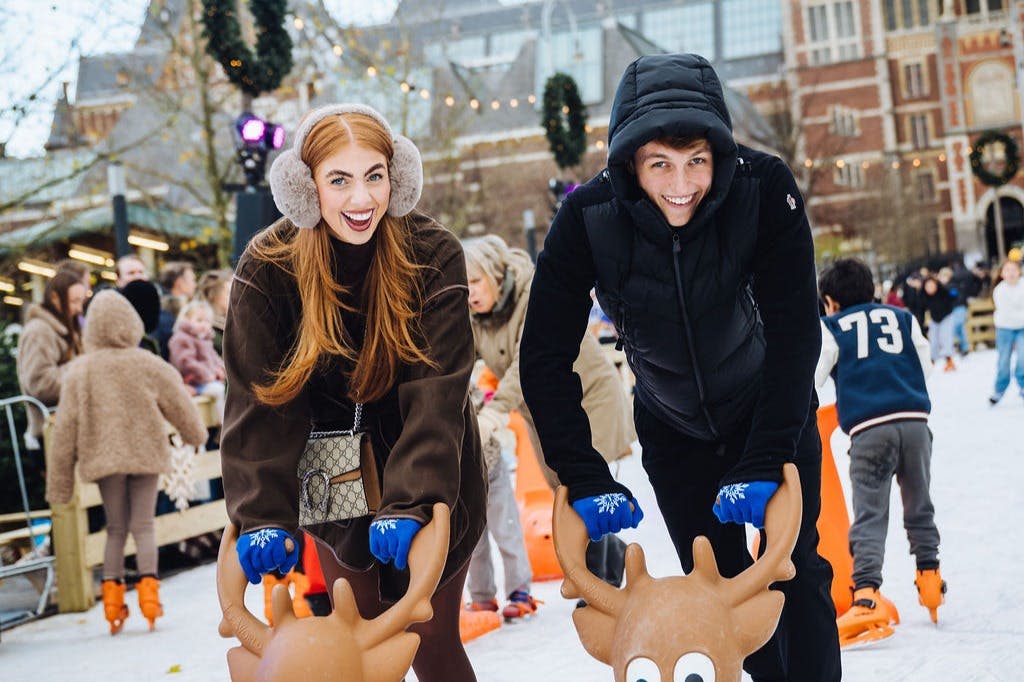 축제 같은 겨울 분위기 속에서 스케이트를 타는 사람들에게 둘러싸인 순록 모양의 지지대를 잡고 두 사람이 신나게 아이스 스케이팅을 하고 있습니다.