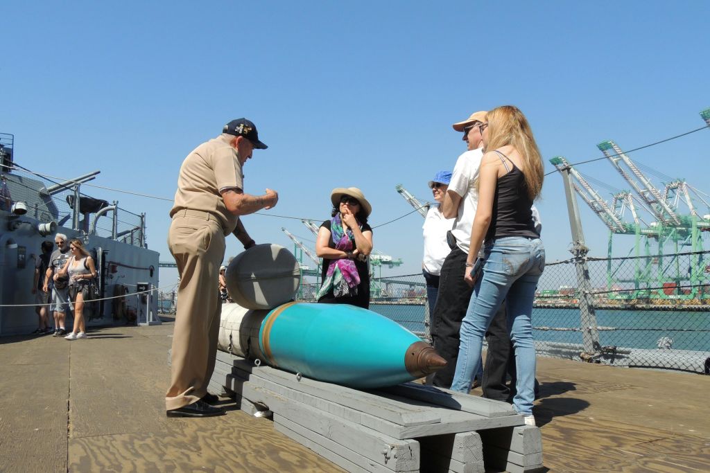 제복을 입은 남성이 커다랗고 푸른 어뢰 같은 물체를 보여주고 있는 선착장의 한 무리. 백그라운드에서 크레인이 보입니다.