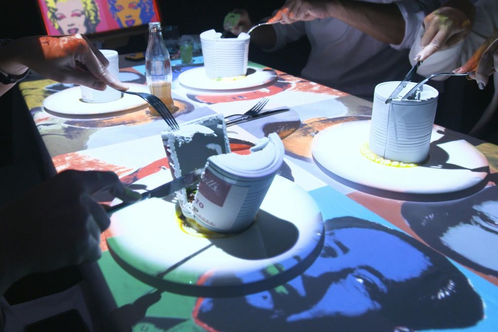 세븐 페인팅스 몰입형 다이닝 쇼에서 게스트들이 식용 워홀 수프 캔을 썰고 있습니다.