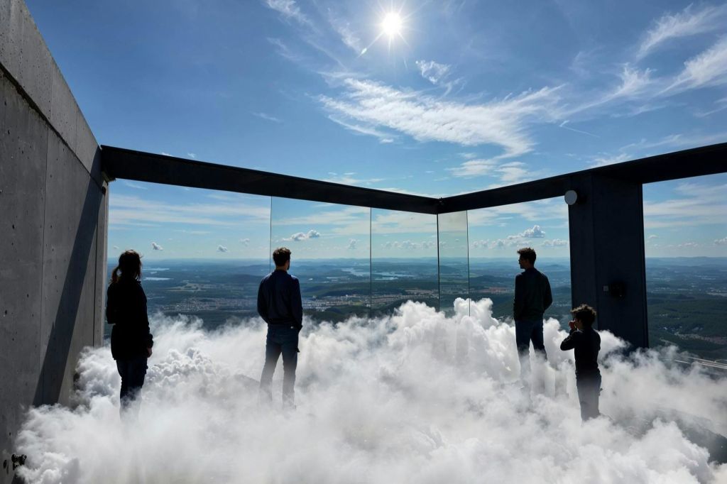 네 사람이 구름으로 둘러싸인 유리 플랫폼에 서서 밝은 태양 아래 광활한 풍경을 내려다보고 있습니다.