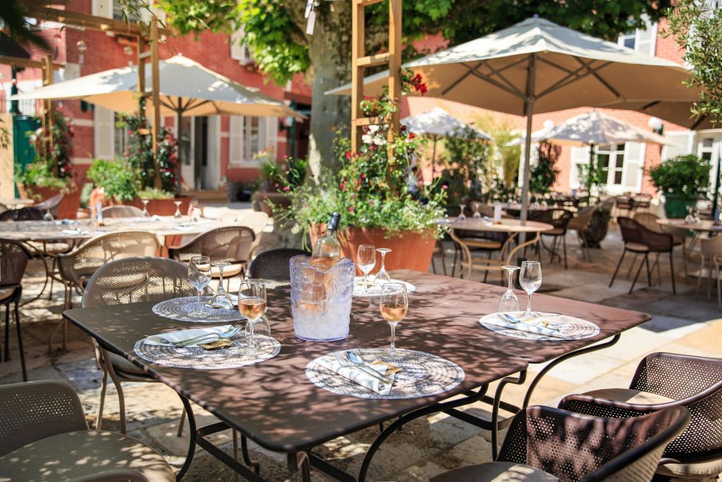커다란 우산 아래 유리 제품, 접시, 냅킨이 놓인 테이블과 식물과 나무로 둘러싸인 야외 레스토랑 파티오.