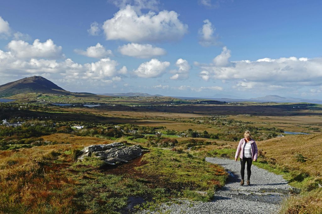 부분적으로 흐린 하늘 아래 광활하고 탁 트인 풍경 속에서 한 여성이 자갈길을 따라 걷고 있습니다.