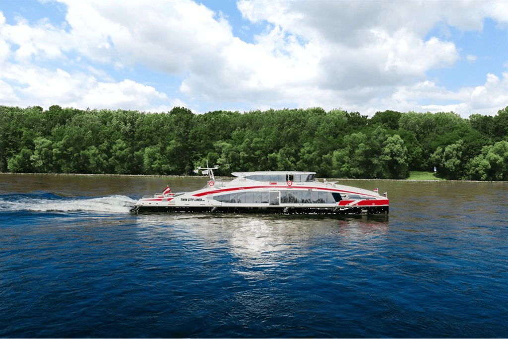 부분적으로 흐린 하늘 아래 가로수가 늘어선 해안을 가로지르는 붉은색과 흰색 디자인의 현대적인 여객 페리.