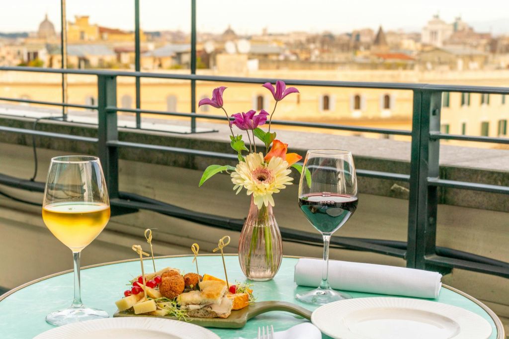 저 멀리 도시 풍경이 내려다보이는 옥상 테이블에 와인잔, 꽃병, 애피타이저가 놓여 있습니다.
