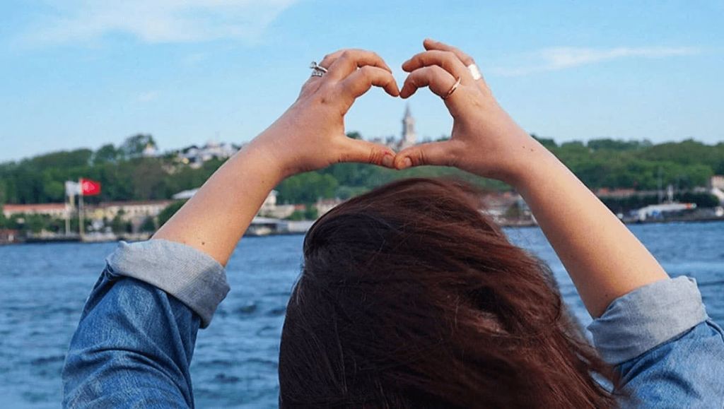 손으로 하트 모양을 만들어 물과 마주하고 있는 사람. 배경에는 건물과 탑이 있는 언덕이 포함됩니다.
