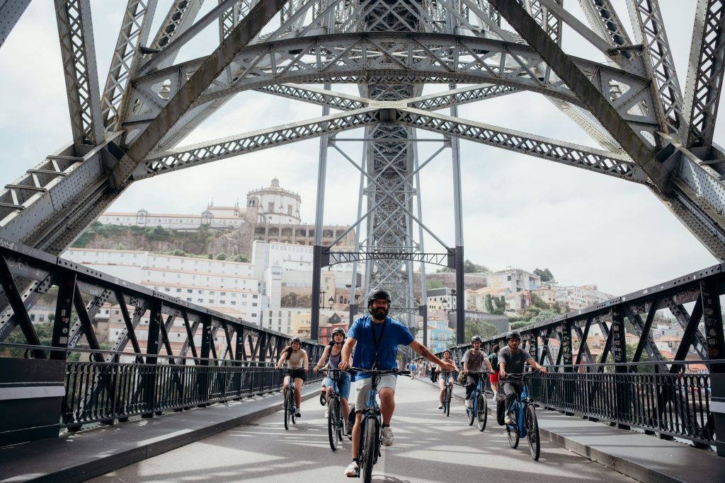 복잡한 금속 구조의 다리를 가로질러 자전거를 타는 사람들로, 도시 풍경이 배경으로 보입니다.