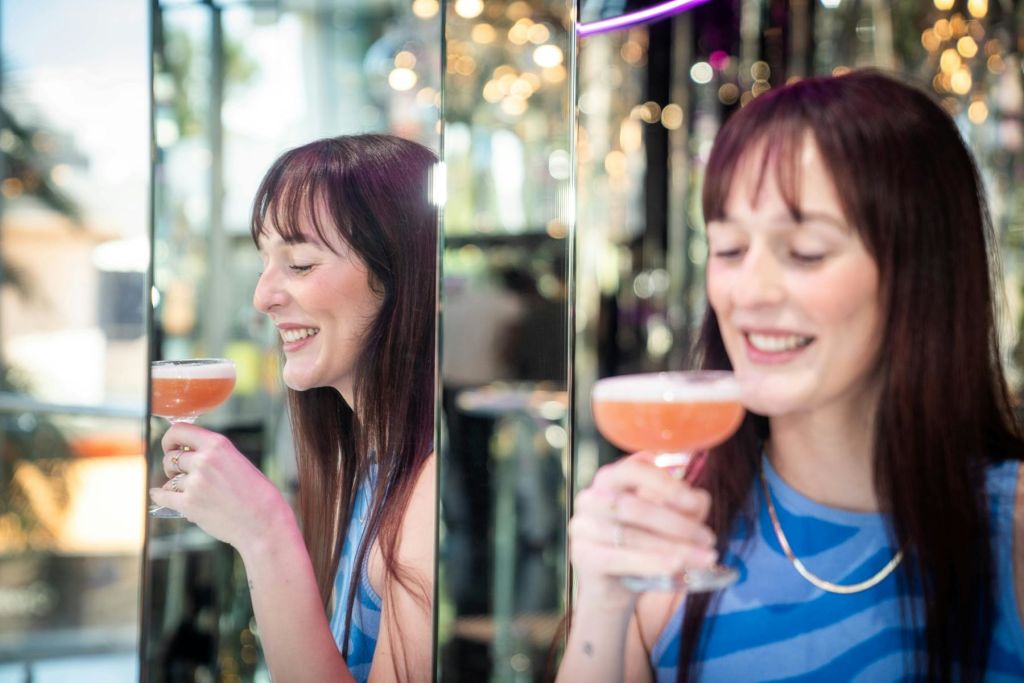 파란색 줄무늬 상의에 긴 머리카락을 한 여성이 칵테일을 들고 미소를 짓고 있습니다. 거울 벽에 그녀의 반사가 보입니다.