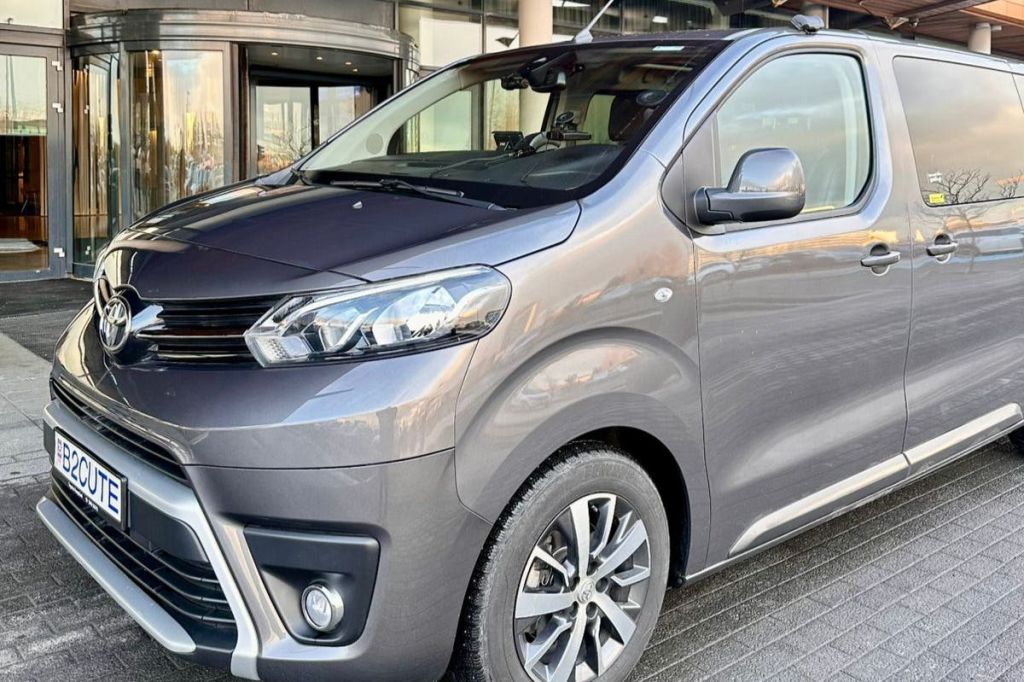 회색 미니밴이 주차되어 있고, 주간 주행등이 켜져 있으며, 앞면과 옆면이 보입니다. 건물 입구가 배경으로 보입니다.