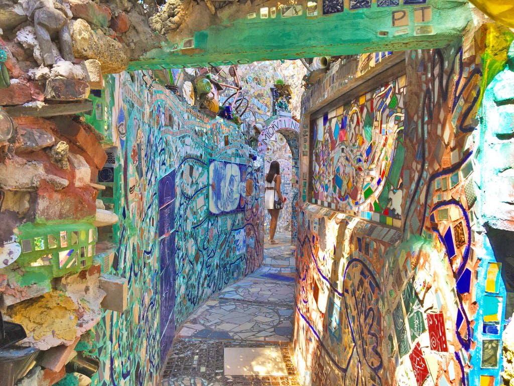 활기찬 모자이크와 조각품으로 장식된 좁은 골목길로, 한 사람이 하얀 가방을 들고 걸어가고 있습니다.
