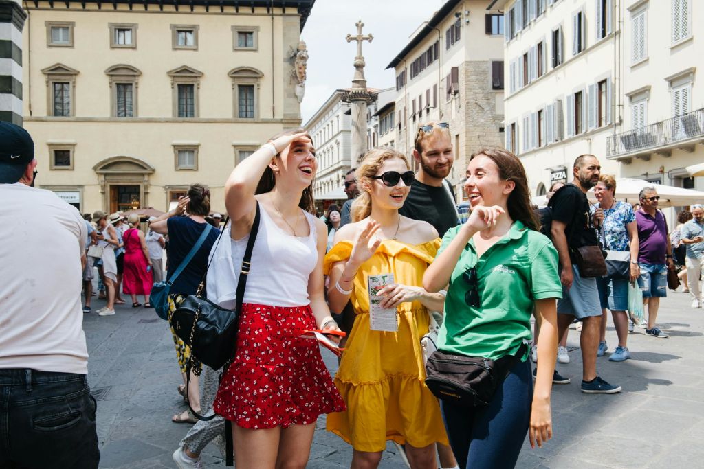 역사적인 건물과 다른 관광객들로 둘러싸인 바쁜 유럽 광장에서 세 명의 여성이 웃으며 이야기하고 있습니다.