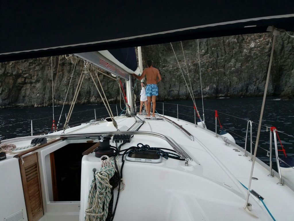 바위 절벽 근처의 요트 갑판에 두 사람이 서 있습니다. 카메라에 더 가까운 것은 셔츠가 없고 파란색 반바지를 입고 있습니다.