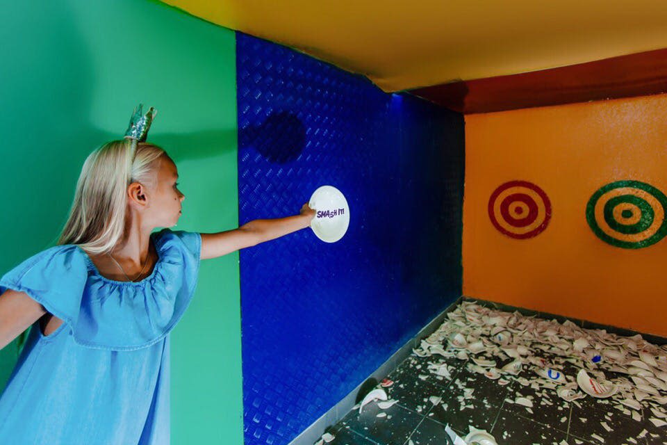 파란 셔츠를 입은 사람이 깨진 접시로 가득 찬 화려한 방에서 하얀 접시를 부수기 위해 준비합니다.