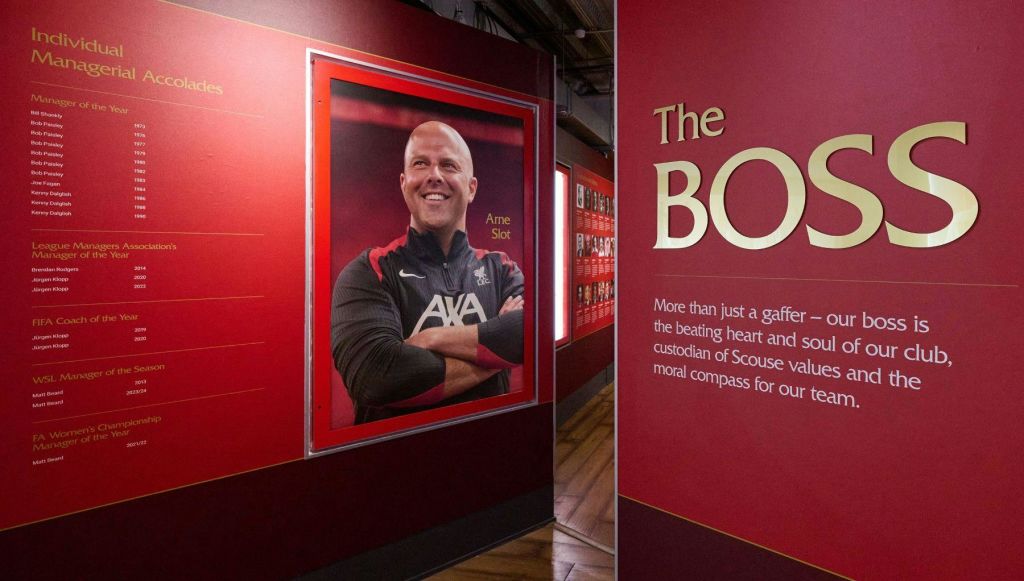 텍스트와 그래픽이 있는 붉은 벽으로 둘러싸인 아르네 슬롯 (Arne Slot) 이라는 스포츠웨어를 입은 웃는 남자의 큰 그림이 있는 박물관 전시관입니다.