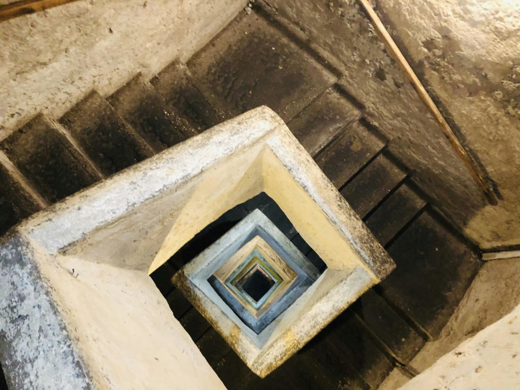 마모된 콘크리트로 만든 정사각형 나선형 계단의 머리 위 모습. 계단은 기하학적 패턴으로 내려갑니다.