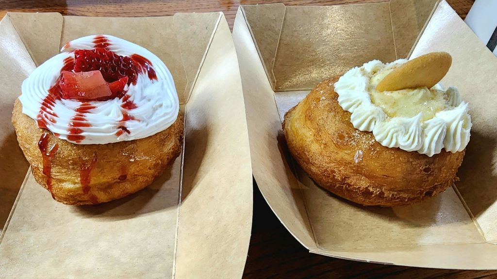 페이스트리 두 개를 종이 트레이에 담고, 하나는 흰색 프로스팅과 빨간색 소스를 얹은 것이고, 다른 하나는 크림과 과일 슬라이스를 얹은 것입니다.
