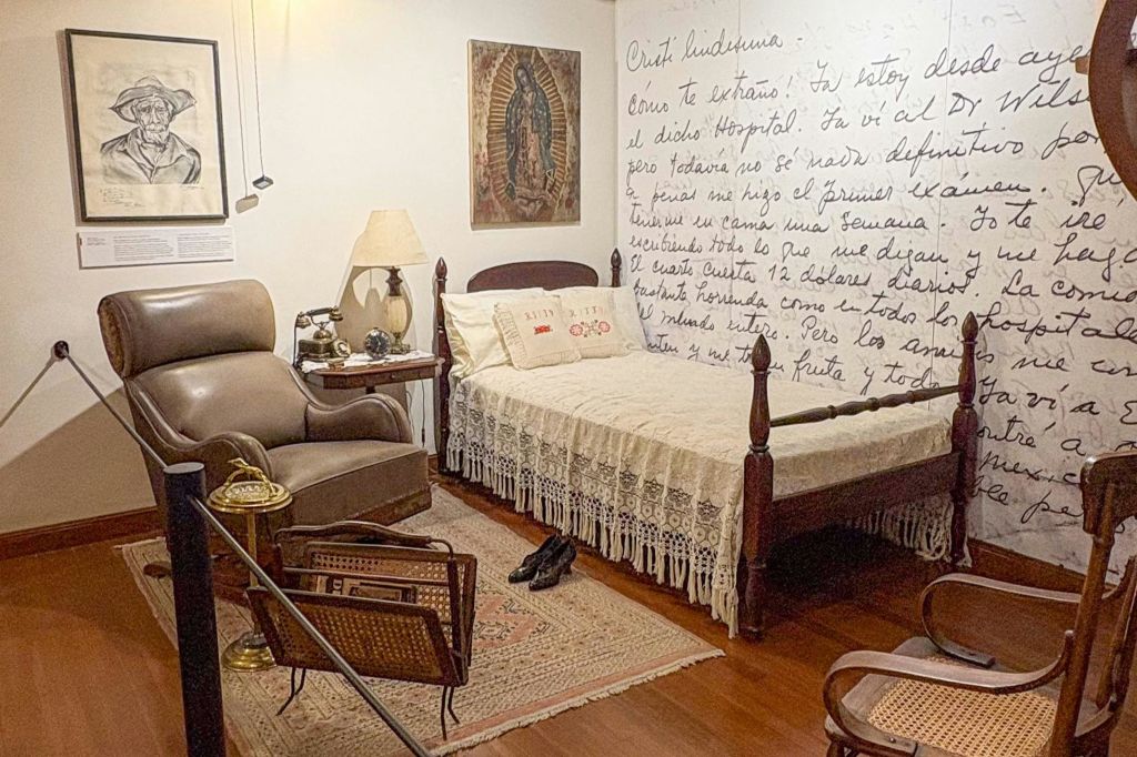 레이스로 덮인 침대, 안락의자, 전등, 액자에 담긴 예술품이 벽에 걸려 있는 아늑한 침실입니다. 한쪽 벽에 손으로 쓴 글씨가 보입니다.